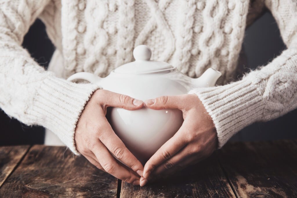 Adult woman hands hug big ceramic teapot with hot drink inside, Adult woman hands hug big ceramic teapot with hot drink inside, making heart sign on it. Close up, wearing cozy warm sweater, everything on aged vintage wooden table